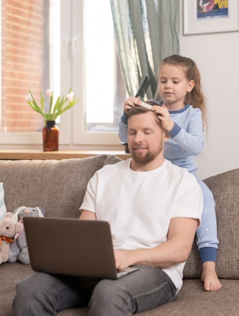 Cute little girl brushing hair of her father with laptop working remotely